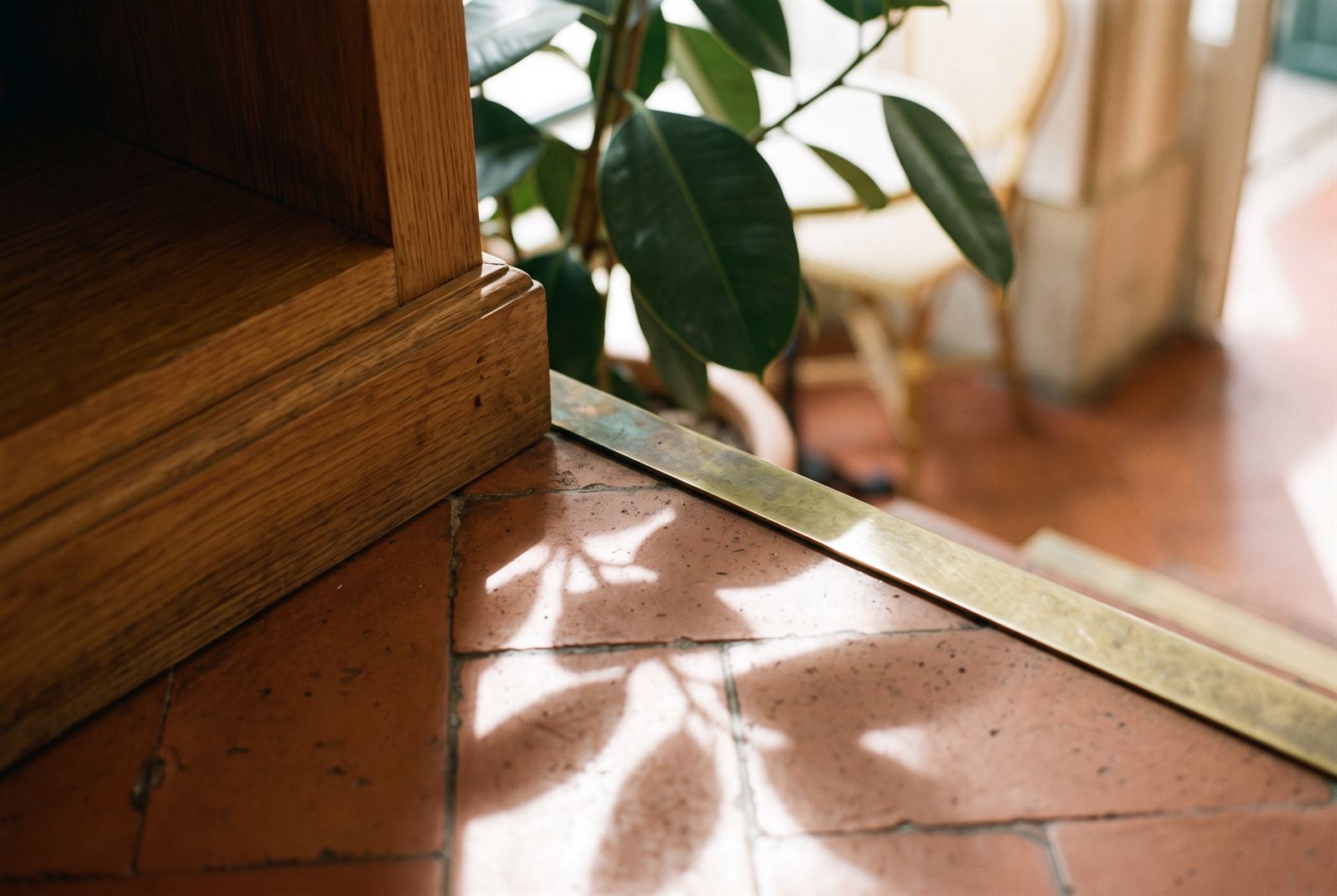 Detail of the oak joinery plinth meeting a brass floor strip set into terracotta tiles, leaf shadows across the junction