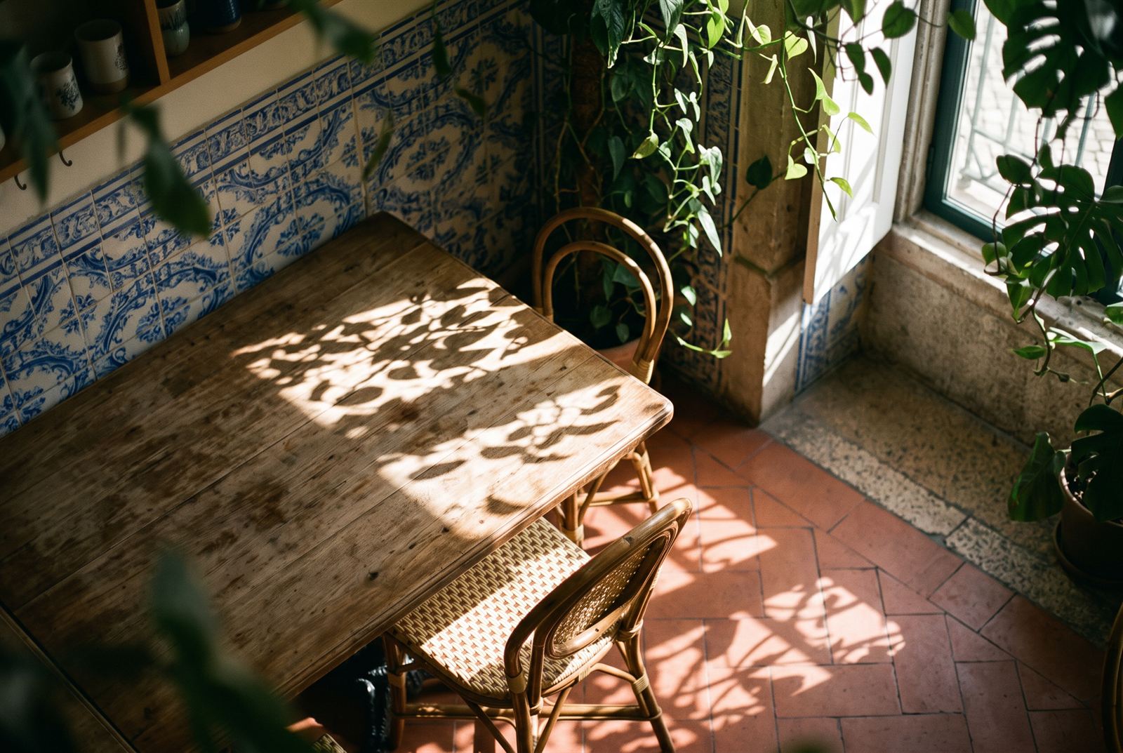 Leaf shadows from the trellis dappling a worn timber tabletop and cane chairs against the blue-and-white tiled wall