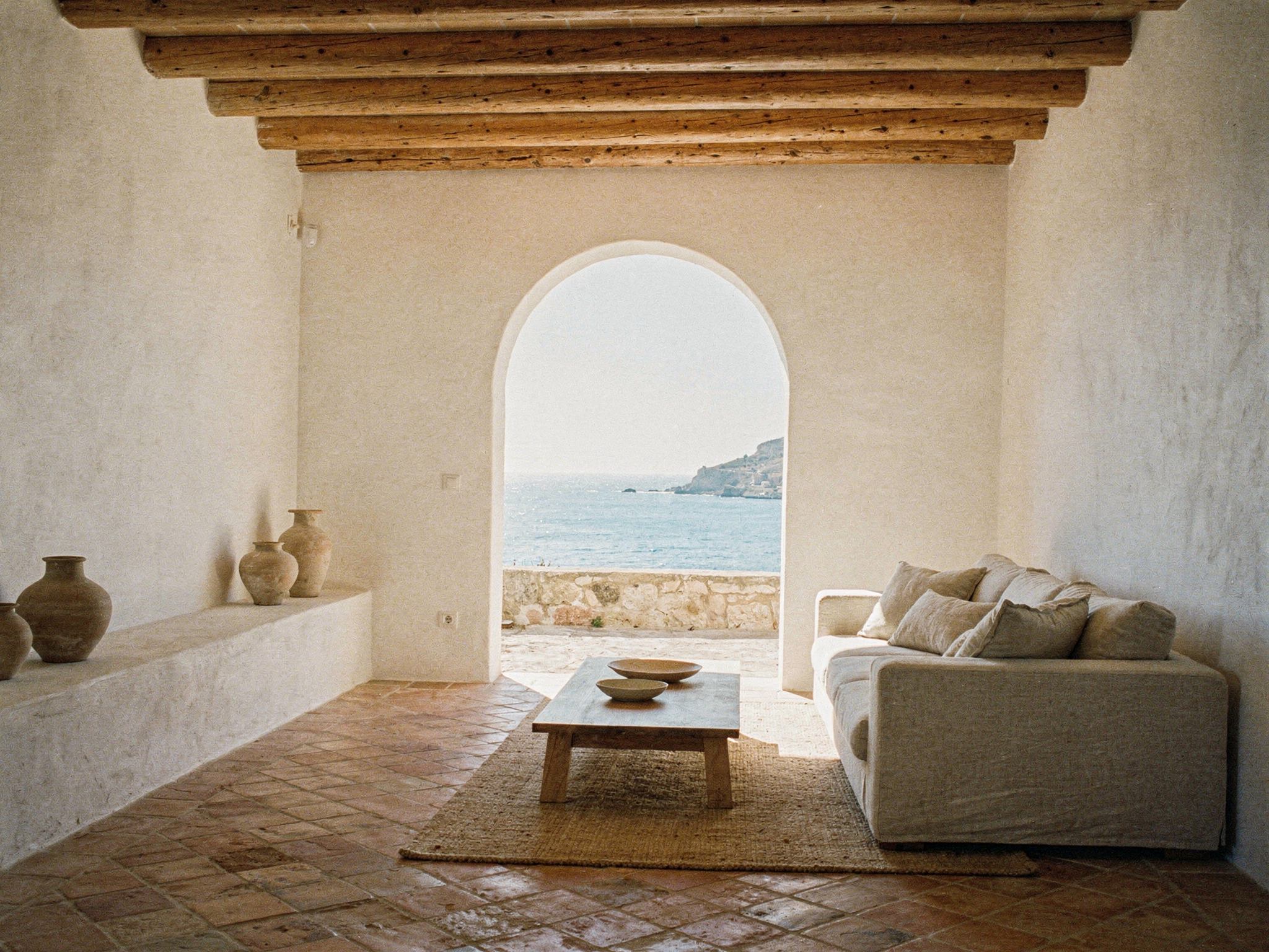 A whitewashed living room with timber beams, terracotta floor and a sea view
