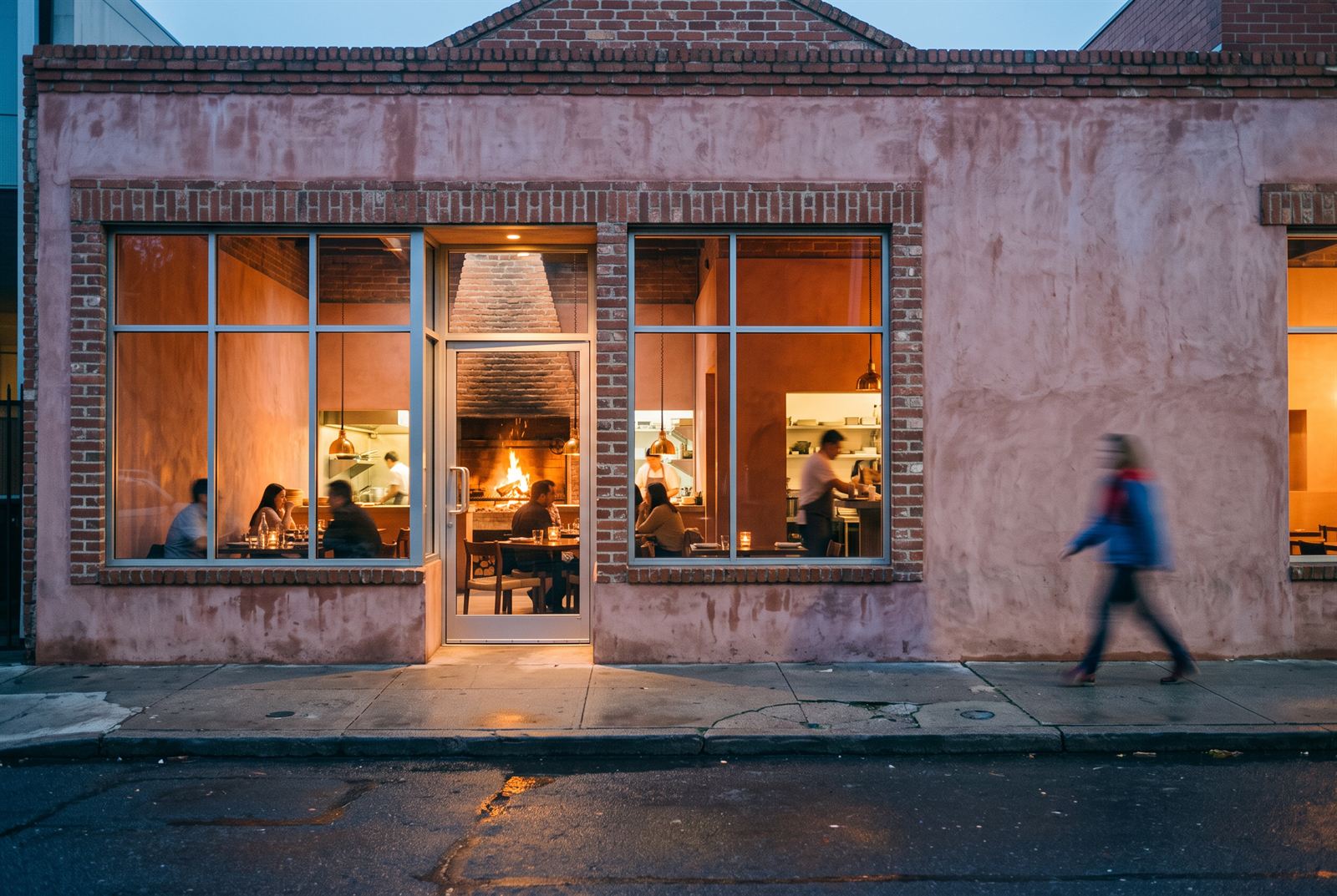 Street elevation at blue hour — the dining room and open kitchen glowing behind brick-edged windows as a passer-by walks the wet footpath
