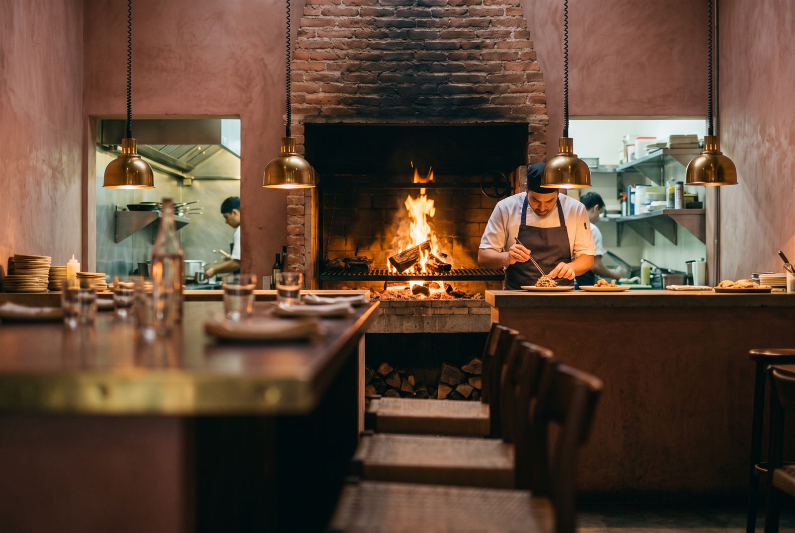 A chef plating at the open-kitchen pass in front of the wood fire burning in the brick hearth