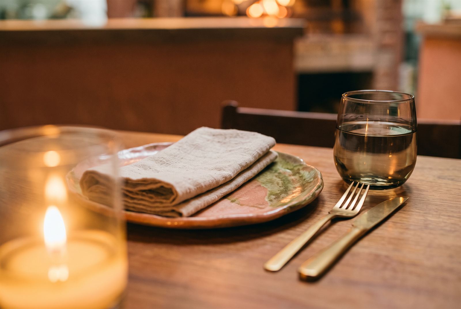 Table detail — a linen napkin folded on a glazed stoneware plate, brass cutlery and a candle, the hearth soft behind