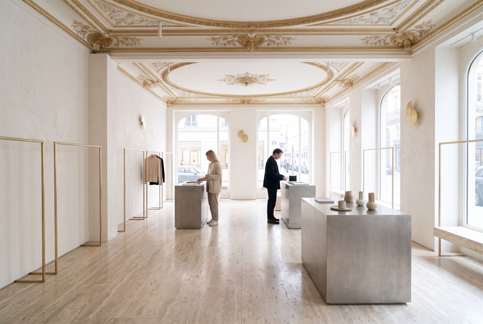 Retail floor with customers browsing at brushed-steel plinths beneath the gilded heritage ceiling