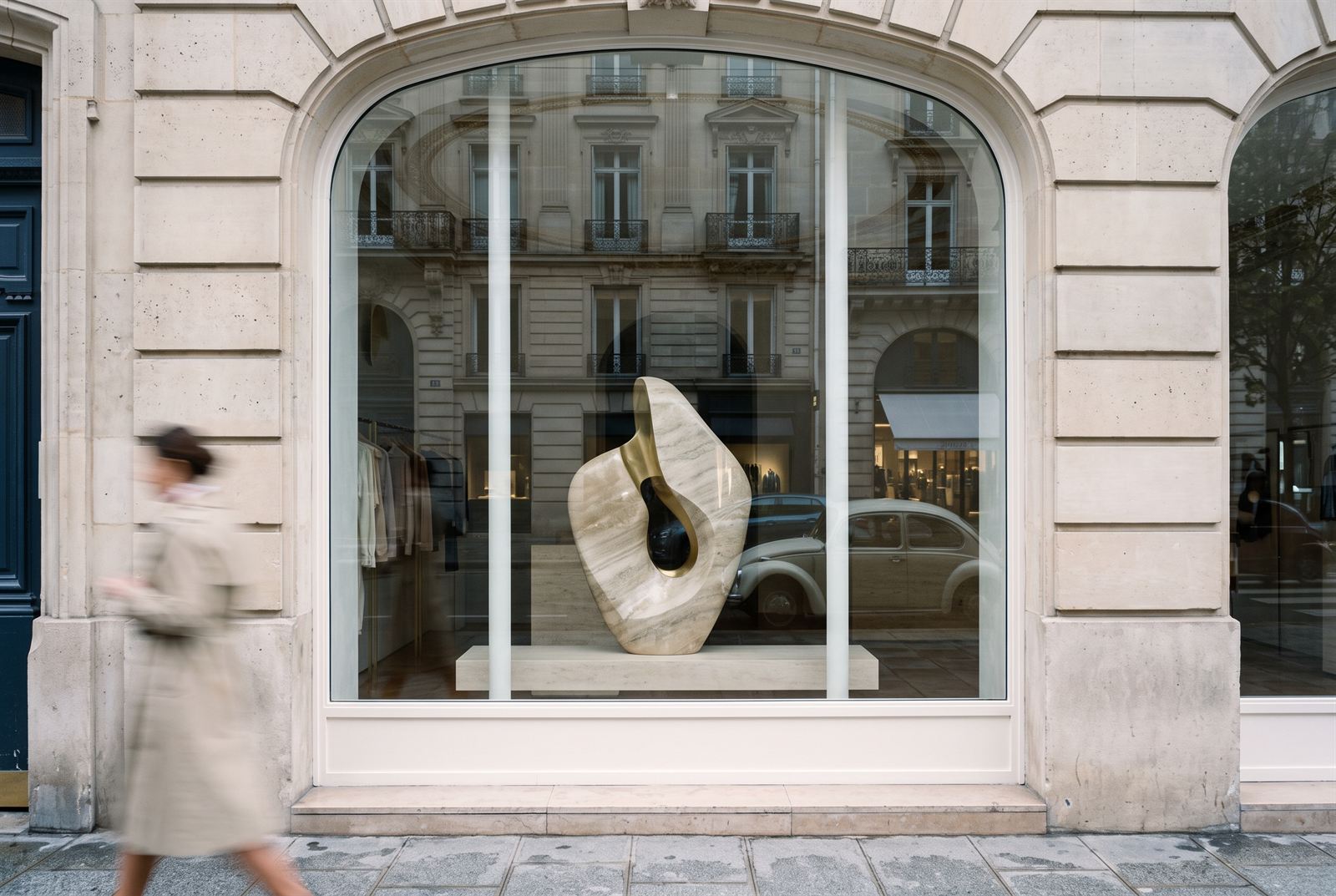 Arched shop window from the street with a stone-and-brass sculpture on a travertine plinth