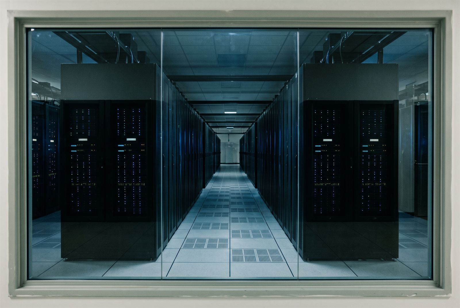 View through a fixed observation window into the server hall, dark racks and status LEDs either side of a perforated-tile aisle